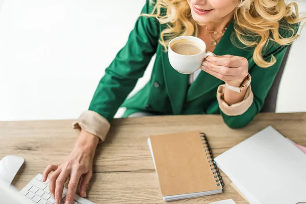 Abgeschnittene Aufnahme einer lächelnden Geschäftsfrau, die einen Desktop-Computer benutzt und am Arbeitsplatz Kaffee trinkt — Stockfoto