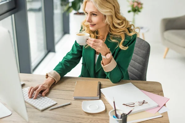 Улыбающаяся деловая женщина за настольным компьютером и пьет кофе на рабочем месте — стоковое фото
