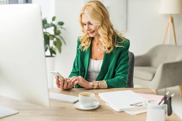 Smiling middle aged businesswoman using smartphone and desktop computer at workplace — Stock Photo