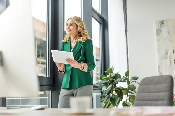 Selektiver Fokus einer lächelnden Geschäftsfrau mittleren Alters, die ein digitales Tablet hält und am Arbeitsplatz wegschaut — Stockfoto