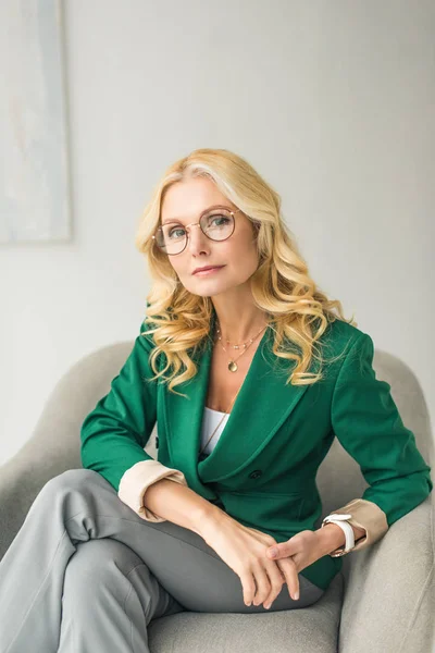 Serious middle aged businesswoman in eyeglasses looking at camera while sitting in armchair — Stock Photo