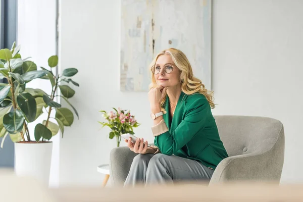 Nachdenkliche Geschäftsfrau mit Brille hält Smartphone in der Hand und schaut weg — Stockfoto