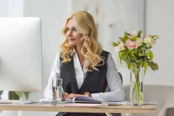 Beautiful middle aged businesswoman taking notes and using desktop computer at workplace — Stock Photo