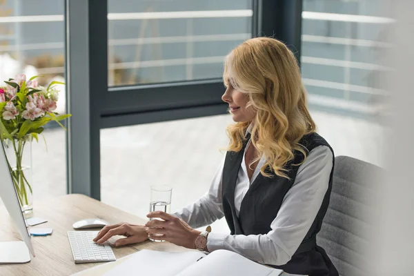 Donna d'affari sorridente che tiene il vetro d'acqua e usa il computer fatto col personal computer sul posto di lavoro — Foto stock