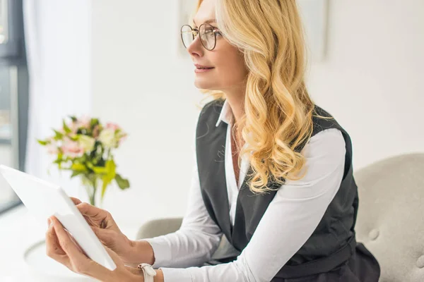 Pensive middle aged businesswoman in eyeglasses using digital tablet and looking away — Stock Photo
