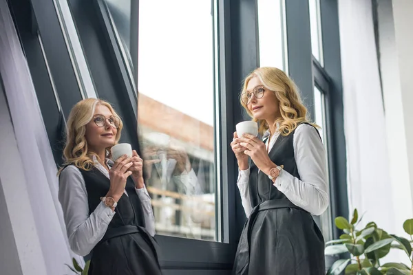 Belle femme d'affaires souriante tenant une tasse de café et regardant la fenêtre avec réflexion — Photo de stock