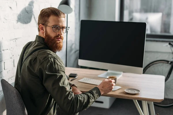 Hombre de negocios barbudo serio sosteniendo una taza de café y mirando a la cámara - foto de stock