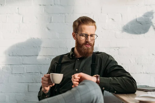 Bel homme d'affaires barbu tenant une tasse de café et vérifiant l'heure — Photo de stock