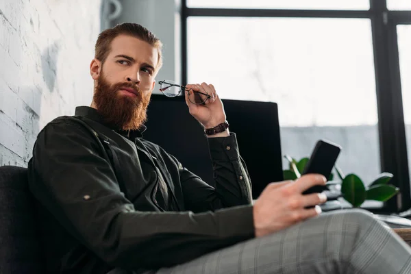 Schöner bärtiger Geschäftsmann sitzt und hält eine Brille mit Smartphone — Stockfoto