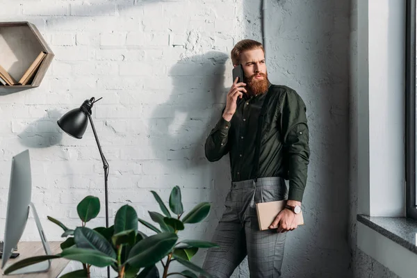 Businessman talking by smartphone, holding book and looking away — Stock Photo