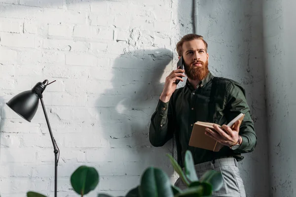 Hombre de negocios hablando por teléfono inteligente, sosteniendo el libro y mirando hacia otro lado - foto de stock