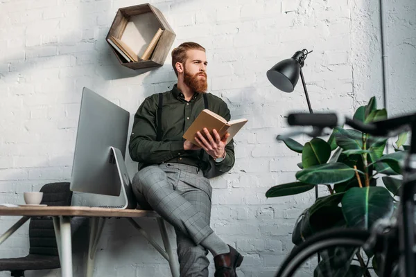 Hombre de negocios apoyado en la mesa, sosteniendo el libro y mirando hacia otro lado - foto de stock