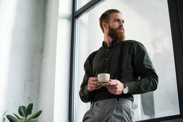 Вид снизу на бизнесмена, стоящего с чашкой кофе и смотрящего в окно — стоковое фото
