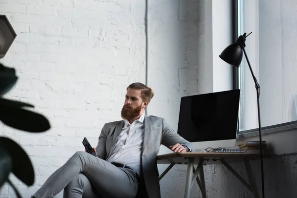 Hombre de negocios sentado en la silla, sosteniendo el teléfono inteligente y mirando hacia otro lado - foto de stock