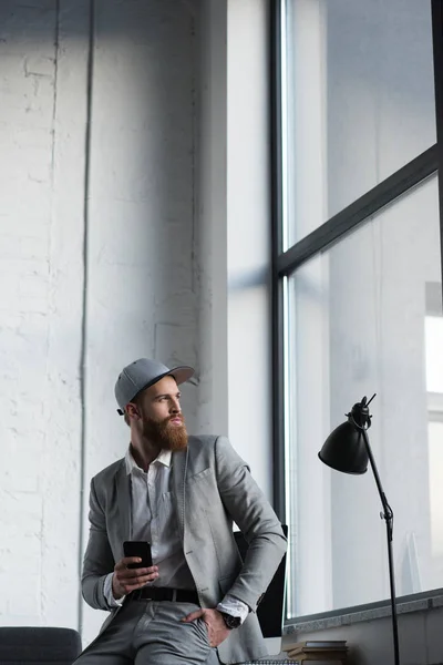 Geschäftsmann mit Baseballkappe hält Smartphone in der Hand und schaut weg — Stockfoto