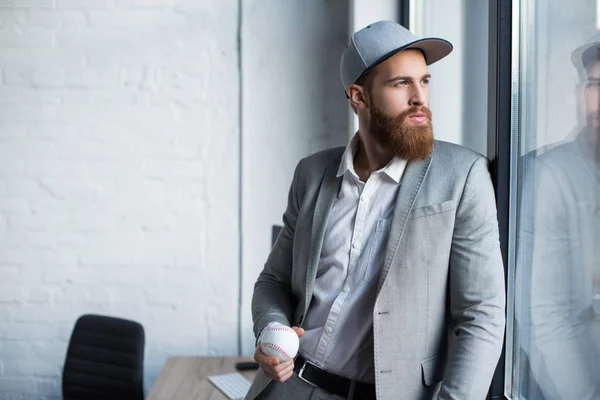 Bärtiger Geschäftsmann mit Baseballkappe blickt auf Fenster und hält Baseballball in der Hand — Stockfoto