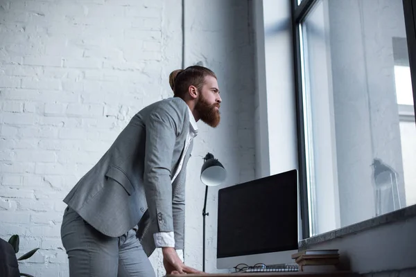 Seitenansicht eines Geschäftsmannes, der sich auf den Tisch lehnt und im Büro ins Fenster schaut — Stockfoto