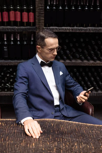 Красивий чоловік у стильному костюмі, використовуючи смартфон у ресторані перед полицями для зберігання вина — стокове фото