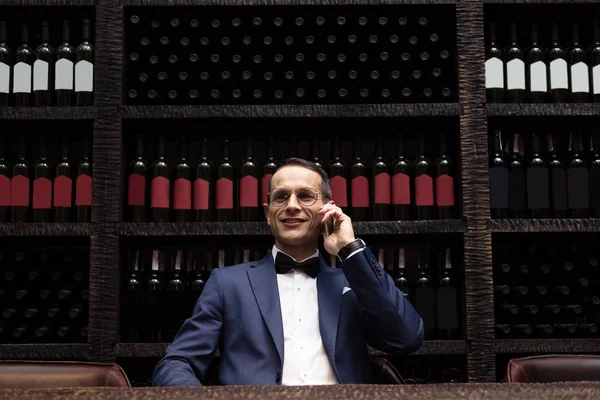 Красивий чоловік у стильному костюмі розмовляє по телефону в ресторані перед полицями для зберігання вина — стокове фото