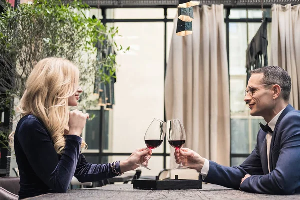 Vista lateral de feliz pareja adulta tintineo vasos de vino en el restaurante - foto de stock
