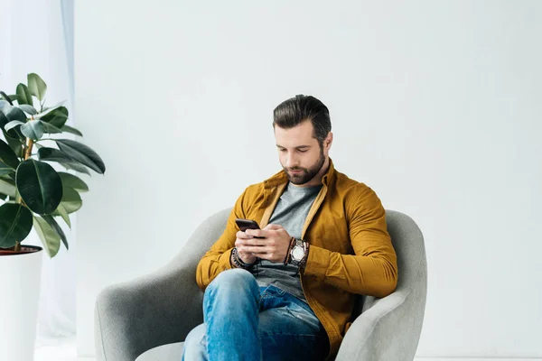Handsome man sitting in armchair and using smartphone — Stock Photo