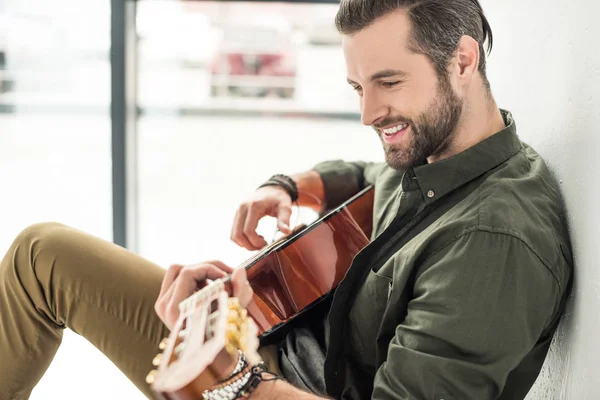 Vista lateral del hombre sonriente guapo tocando la guitarra acústica - foto de stock