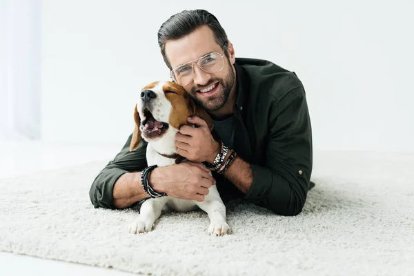 Sonriente hombre guapo acostado en la alfombra con lindo beagle y mirando a la cámara - foto de stock