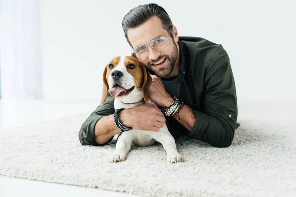 Happy handsome man lying on carpet with dog and looking at camera — Stock Photo