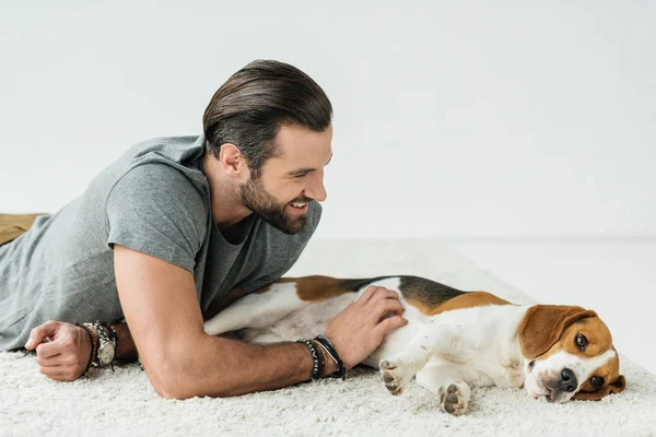 Sonriente hombre guapo acostado con lindo beagle en la alfombra - foto de stock