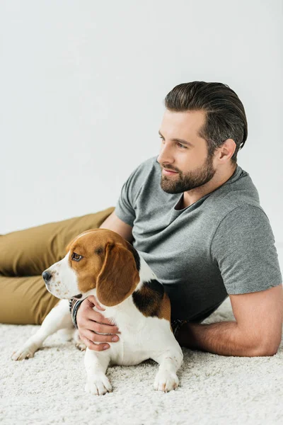 Handsome man lying with cute beagle on carpet and looking away — Stock Photo