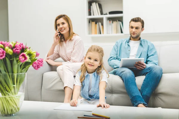 Hija dibujo imagen mientras los padres utilizando dispositivos digitales en casa - foto de stock