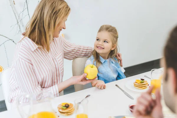 Madre dando manzana a la hija durante el desayuno en casa - foto de stock