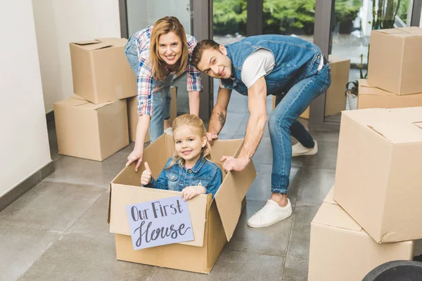 Genitori sorridenti che giocano con la piccola figlia con la nostra prima casa in scatola di cartone nella nuova casa — Foto stock