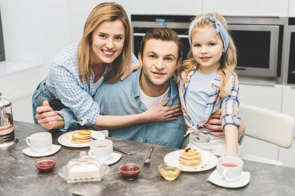 Retrato de padres sonrientes e hija mirando a la cámara durante el desayuno en casa - foto de stock