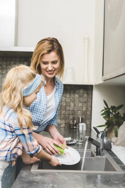 Side view of little daughter helping mother to wash dishes after dinner in kitchen — Stock Photo