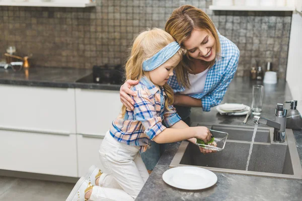 Side view of little daughter helping mother to wash dishes after dinner in kitchen — Stock Photo