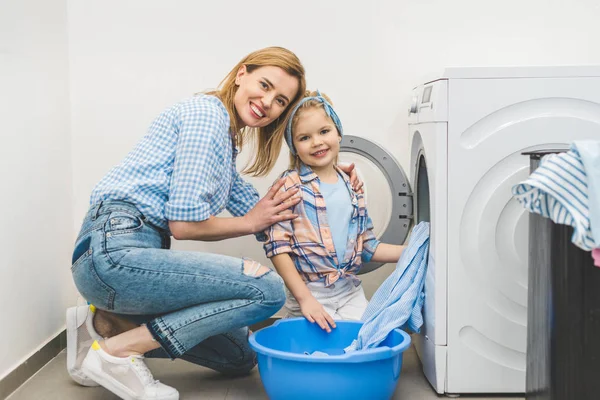 Мати і дочка дивляться на камеру, вкладаючи одяг в пральну машину вдома — стокове фото