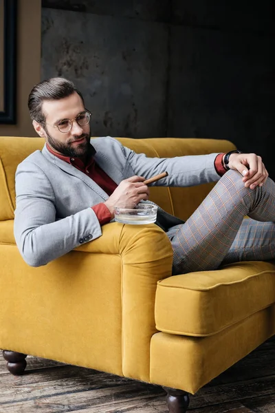 Стильний красивий чоловік з сигарою і попільничкою сидить на дивані — Stock Photo