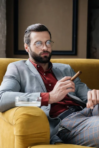 Homem elegante com charuto e cinzeiro sentado no sofá — Fotografia de Stock