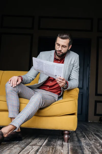 Bonito homem na moda terno ler jornal e sentado no sofá — Fotografia de Stock