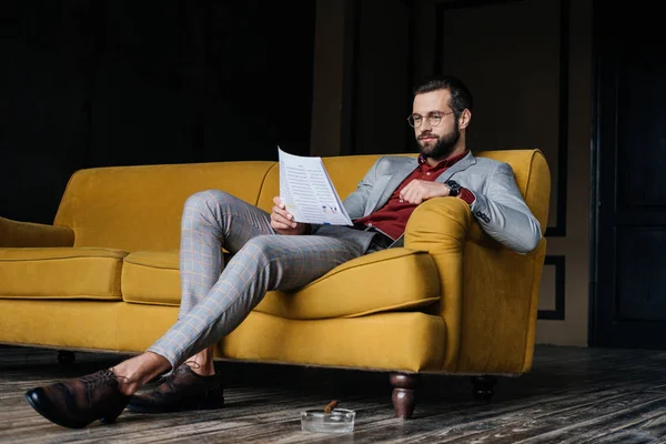 Élégant homme lisant le journal et assis sur le canapé, cendrier avec cigare sur le sol — Photo de stock