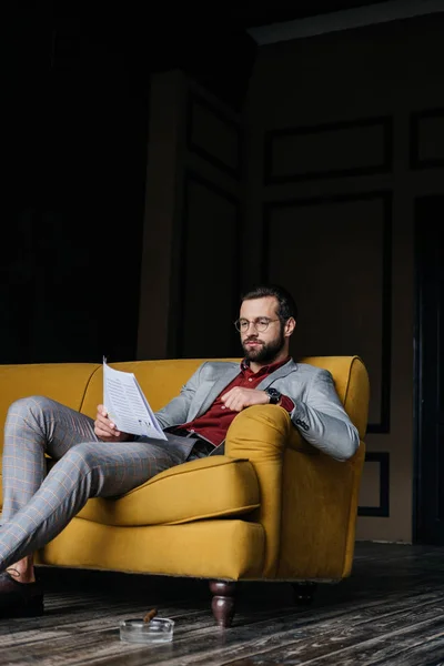 Bel homme lisant le journal et assis sur le canapé, cendrier avec cigare sur le sol — Photo de stock
