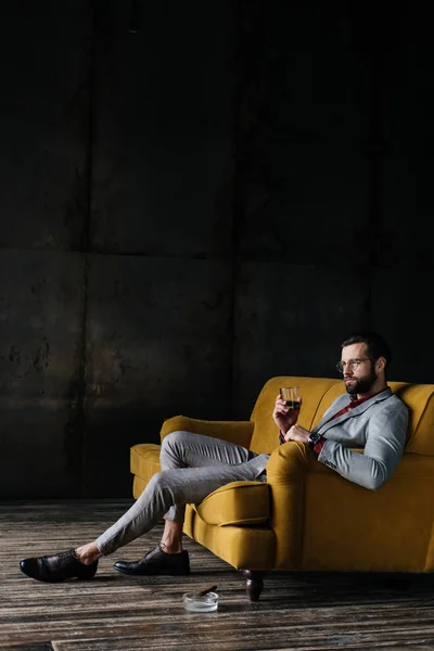 Hombre de moda con vaso de whisky sentado en el sofá con cigarro en el cenicero en el suelo - foto de stock