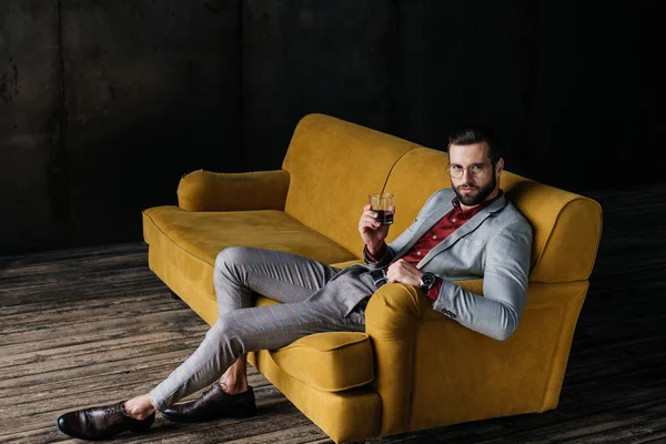 Elegante hombre guapo sosteniendo vaso de coñac y sentado en el sofá amarillo - foto de stock