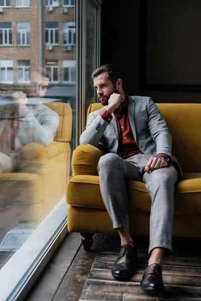 Stylish thoughtful man sitting on yellow sofa at window — Stock Photo