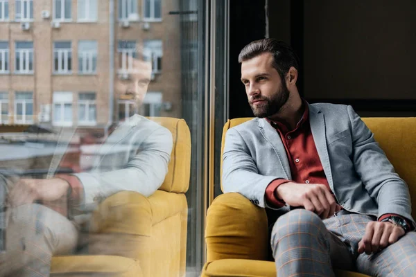 Красивый бородатый мужчина сидит на жёлтом диване у окна с отражением — стоковое фото