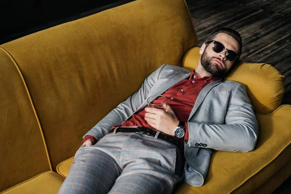 Stylish drunk man in sunglasses with glass of cognac lying on yellow sofa — Stock Photo