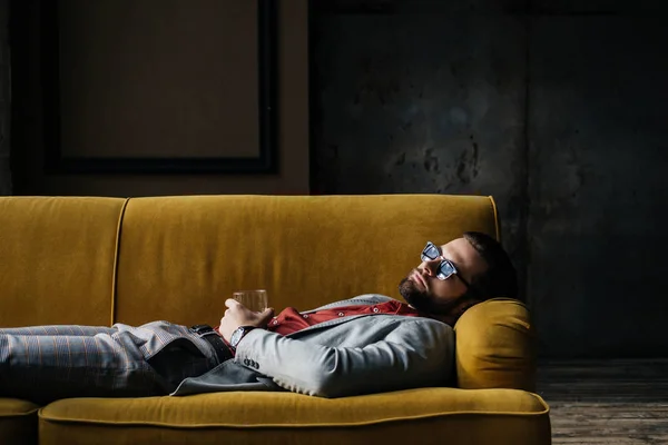 Homme ivre en lunettes de soleil avec verre de cognac couché sur un canapé jaune à l'intérieur du grenier — Photo de stock