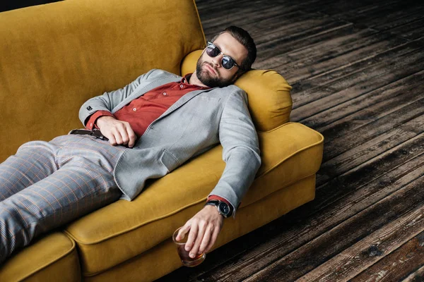 Hombre borracho con estilo en gafas de sol sosteniendo vaso de whisky y acostado en un sofá amarillo - foto de stock