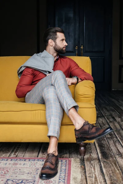 Hombre de moda en traje y zapatos brogue sentado en el sofá amarillo en el interior del loft - foto de stock